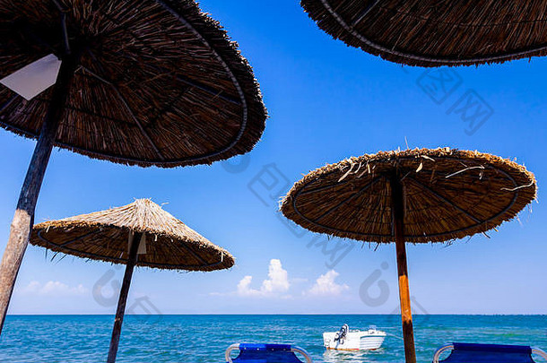 在空荡荡的公共海滩上，沿着沙质海岸线可以看到茅草遮阳棚、雨伞和躺椅。小型突堤船停泊在海滩附近。