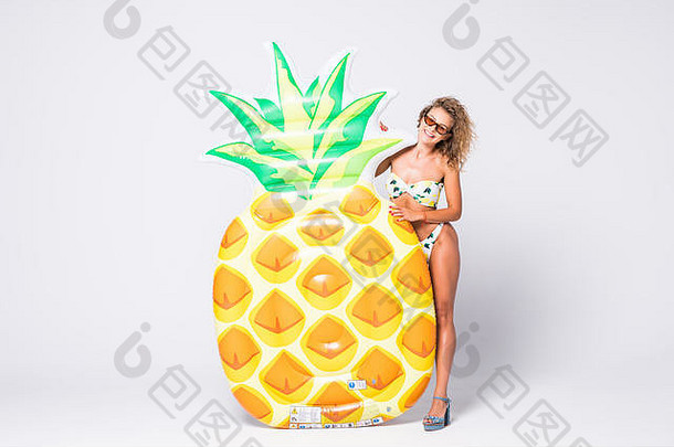 一位身穿比基尼的年轻女子站在白色背景下，带着充气菠萝摆姿势的全长肖像