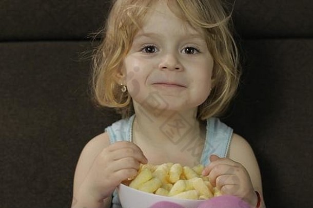 漂亮的金发小女孩坐在家里的沙发上，吃着玉米泡芙。可爱的孩子喜欢吃爆米花。在…内儿童餐
