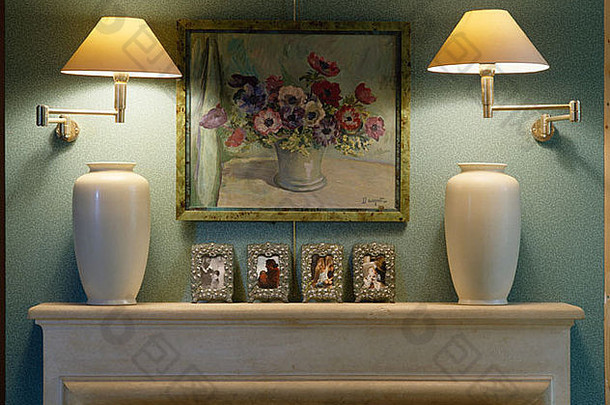 壁炉台上方的花卉画两侧的照明壁灯特写，带有两个白色花瓶和相框照片
