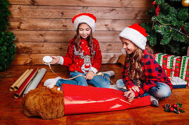 戴着<strong>圣诞</strong>帽的快乐有趣的小姐妹们在<strong>圣诞</strong>树旁打包礼物