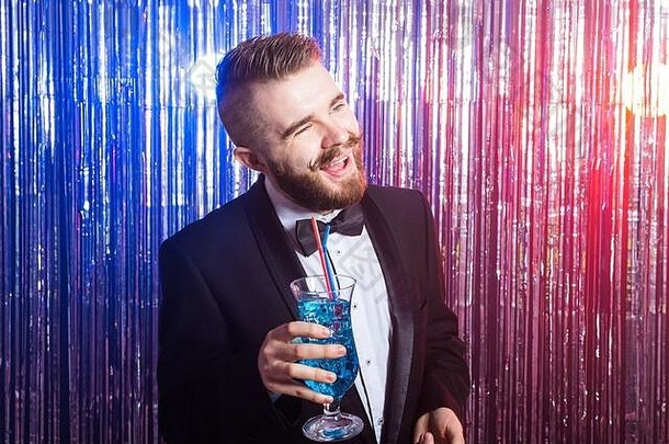 俱乐部派对和假日概念-优雅快乐帅哥的肖像，穿着昂贵的西装，在闪亮的背景上拿着蓝色鸡尾酒。