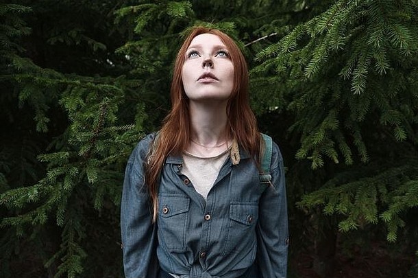 一个穿着牛仔衫、蓝眼睛的红发女孩站直了身子，抬起头，把头靠在冷杉树的背景上