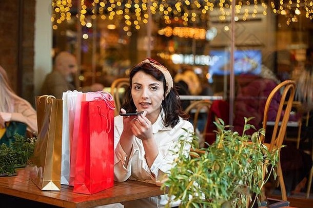 迷人的美女在购物后坐在咖啡馆里，手持购物袋在智能手机上启动虚拟数字语音助手，并与之交谈