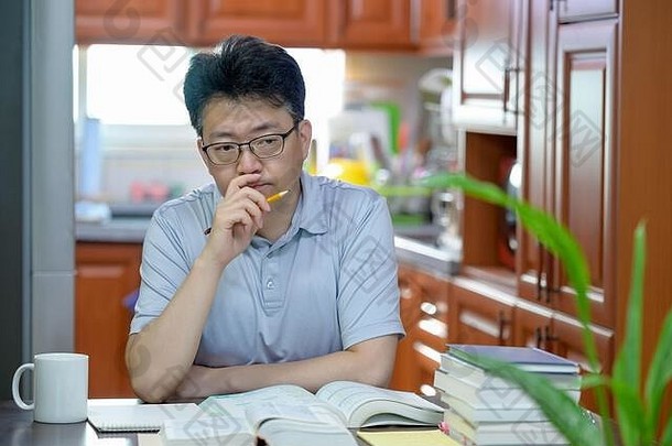 一位亚洲中年男子坐在家里的书桌旁，读书和学习。