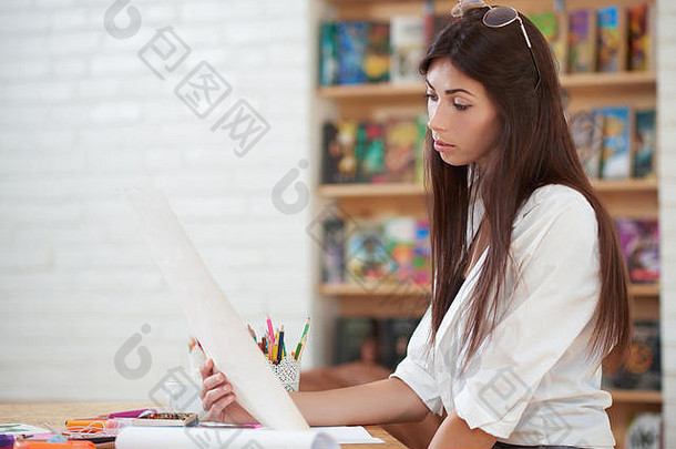 女艺术家思考并看着她的插图。专业画家手里拿着一张纸，坐在桌子旁，手里拿着不同的画家用品。艺术工作室里的美女。