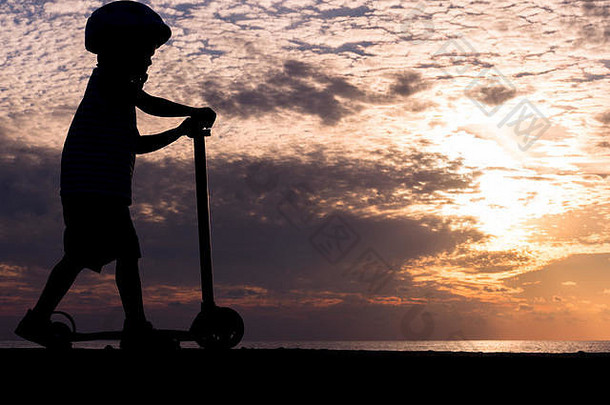 轮廓男孩穿头盔骑踏板车背景海日落