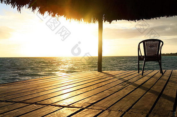 日落或日出时海滩上的异国情调的木制露台和椅子。背景是一个海滨，有座椅、休息亭、棕榈树和蓝色的海水