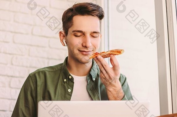饥肠辘辘的时髦男自由撰稿人一边闻着，一边拿着一片美味的意大利披萨，一边在窗台上远程学习。快乐商人<strong>吃外卖</strong>快餐