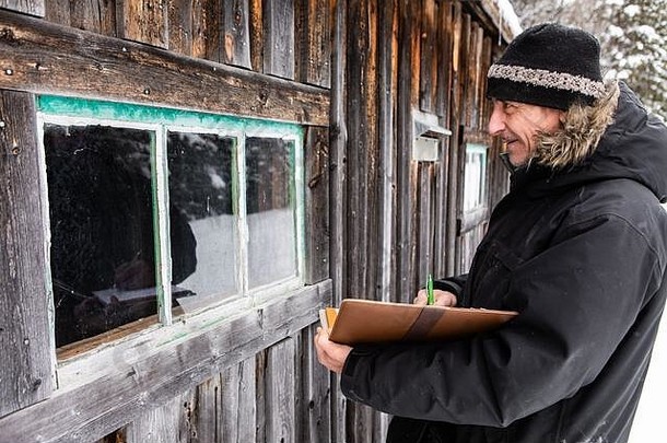 中年家政督察在一次户外检查中，在一本皮封面的笔记本上记笔记。检查旧木屋的窗户