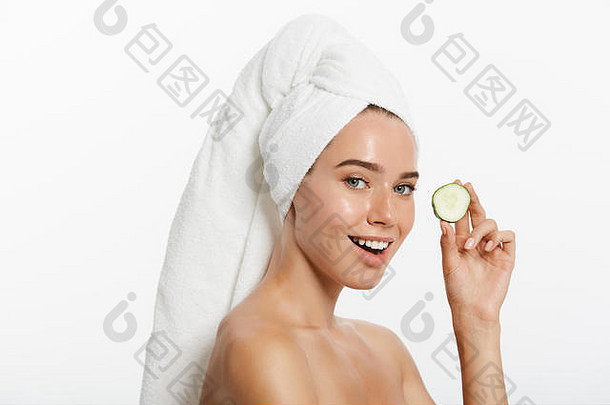 一位微笑的半裸美女的特写美女肖像，用毛巾裹着头发，拿着黄瓜片，看着隔离在白色背景上的摄像机