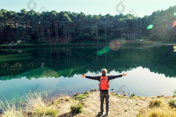 背景：年轻的旅游男子背着背包，双臂举向天空，站在森林湖附近庆祝自由。探索世界。徒步旅行