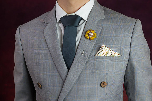 身着灰色西装、格子花纹、蓝色领带、花朵胸针和点图案手帕的男士，特写