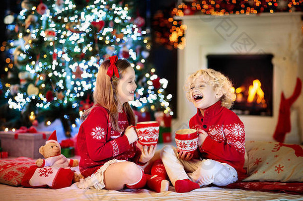 孩子在家里的圣诞树下。穿着带有圣诞节装饰的针织毛衣的小男孩和小女孩喝热巧克力。一家人带着孩子庆祝冬天