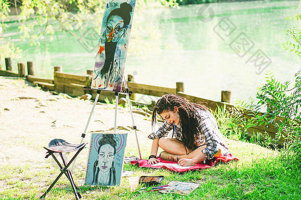年轻的艺术家女孩在湖边的公园里画下一幅画的草稿——一位留着可怕长发的女画家在城市户外从事她的艺术创作