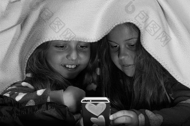 女朋友在毯子下玩手机。孩子们穿着黑色背景的红色睡衣躺在床上。儿童与技术概念。有着好奇面孔的女孩。儿童睡衣派对。