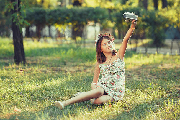 这个小女孩坐在公园的草地上，被一架照相机所扮演。自拍和拍摄世界各地