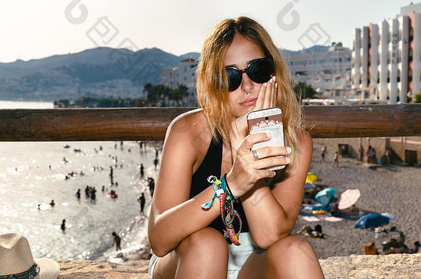一位戴着太阳镜的年轻白人妇女坐在一座防波堤的石墙前，海滩上挤满了游客。她正在看她的智能手机，上面有t
