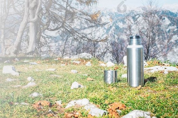 在美丽山峦的背景下，绿草上放着金属保温瓶和热茶。