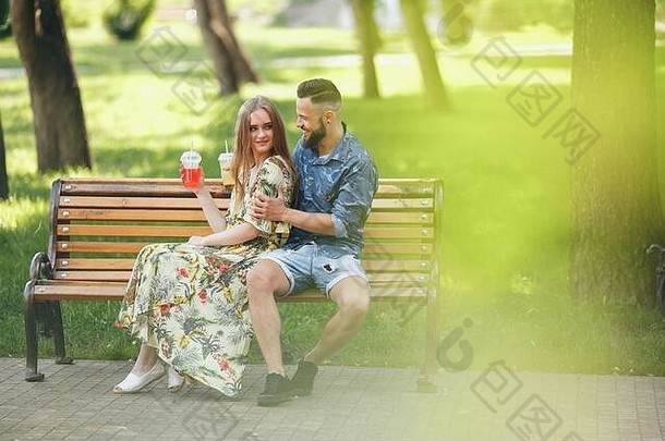 阳光明媚的夏日里，一对年轻时尚的情侣坐在长椅上，在城市公园里休息，喝着饮料