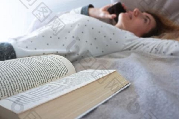 一个女孩穿着睡衣呆在卧室里，一边用智能手机打电话，一边在床上<strong>翻开书本</strong>——在家里放松地呆在家里