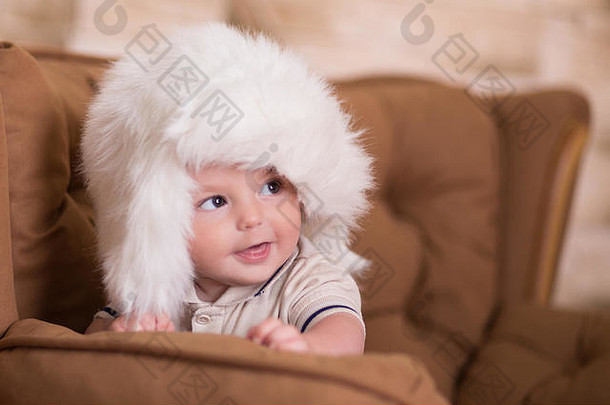 快乐的新生婴儿面颊粉红色的在黑格复古休闲风格的棕色沙发沙发沙发上摆姿势，沙发上戴着白色毛皮<strong>冬季保暖</strong>帽。