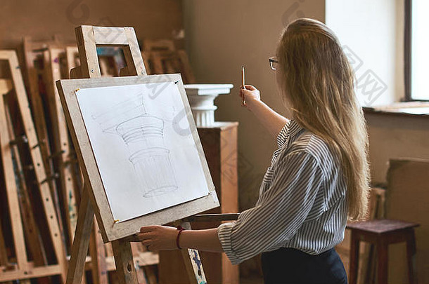 年轻漂亮的女学生正在木画架上用铅笔画一个多立克柱头，这是她的大学作业