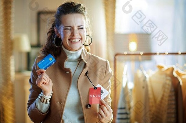 一位40岁的女士穿着白色毛衣和裙子，兴奋地在现代时装店里尝试着米色外套，上面有红色的销售价格标签，手里拿着蓝色的。
