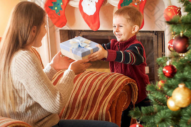 微笑的小男孩坐在客厅的大扶手椅上，给他妈妈送圣诞礼物盒的画像