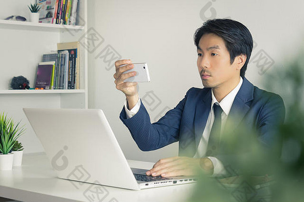 亚洲商人在办公室的笔记本电脑显示器前用智能手机拍照或自拍。亚洲商人放松时间