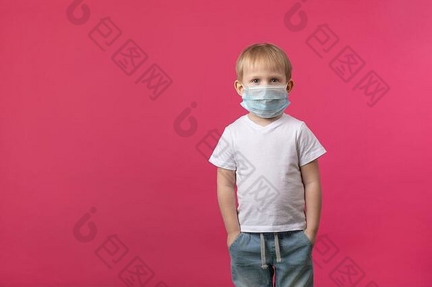 一个金发碧眼的欧洲男孩站在一个冠状病毒的医用面具里看着摄像机。工作室在粉色背景上拍摄，地点为fo