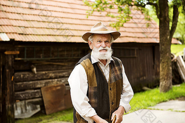 一位留着胡子戴着帽子的老农站在他的农舍旁