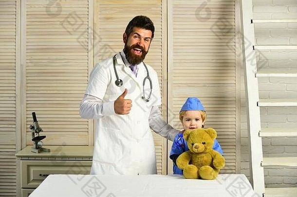 兽医和助手抱着玩具宠物，竖起大拇指。保健和儿童概念。留着胡子的男人和男孩在木制背景上玩泰迪熊。爸爸和孩子开心地扮演医生