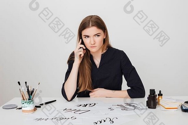 一张女士坐在白色办公桌旁，一边用手机聊天，一边认真地向一边看的照片