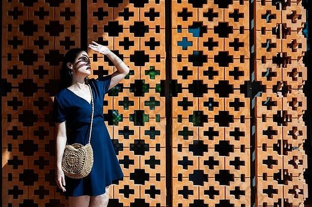 身穿深蓝色连衣裙的长发女人站在砖墙前，举手保护眼睛不受阳光照射。