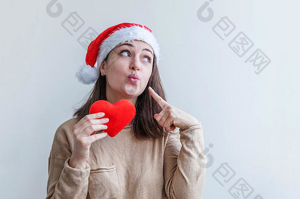 戴着红色圣诞老人帽子的美丽女孩，手拿着红色的心，在白色背景上显得快乐和兴奋。年轻女子肖像，真实情感。圣诞快乐，新年快乐。