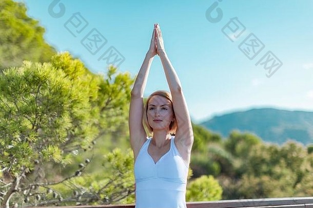 一幅严肃的年轻女子在户外练习瑜伽的画像，双手高举在蓝天和绿树的映衬下
