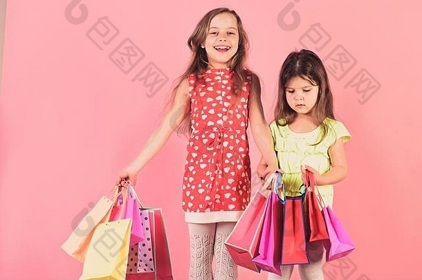 购物狂、童年和时尚概念。女士买衣服或礼物。脸上带着快乐和好奇的女孩们拿着粉色背景的购物袋。女学生拿着粉红色、红色和黄色的小包