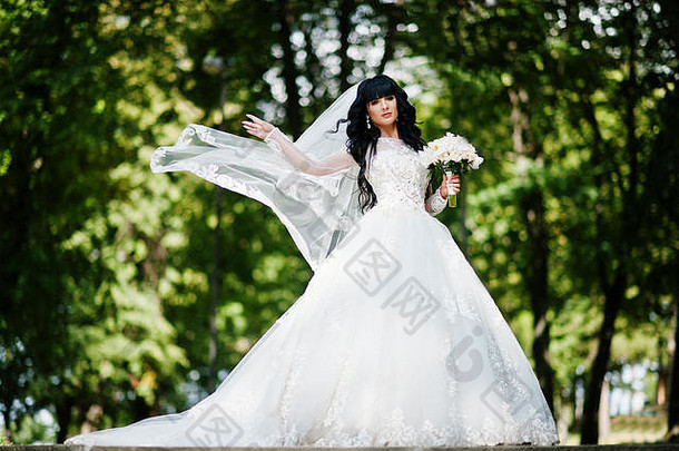 美丽的卷曲的深褐色新娘，手戴面纱和婚礼花束，在公园里摆姿势。