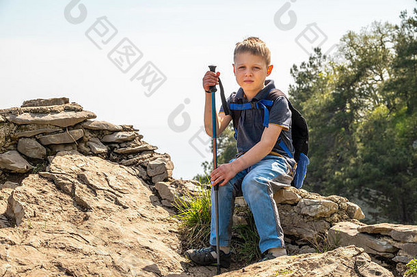 一个带着登山杖和背包的男孩在绿色森林中的山顶上休息。山里春夏晴朗