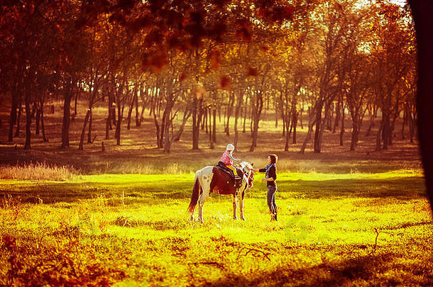 小女孩骑在马背上，妈妈在附近散步