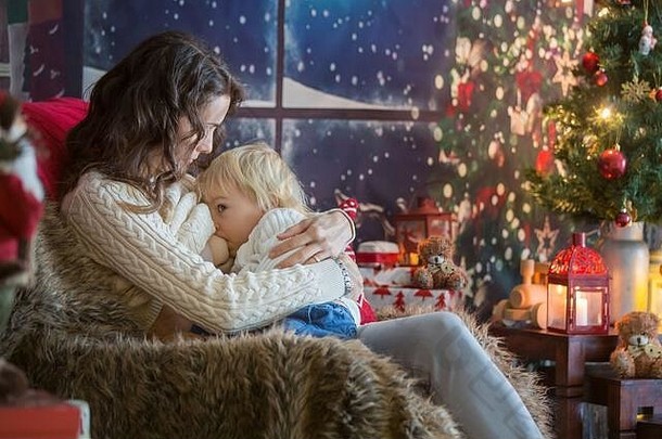美丽的年轻妈妈和金发的蹒跚学步的男孩，坐在舒适的客厅里，为<strong>圣诞</strong>节装饰，哺乳，拥抱和亲吻