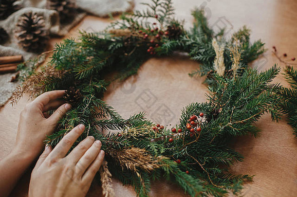 手拿着圣诞花环，上面有冷杉枝、浆果、松果和线，剪刀放在乡村的木桌上。圣诞花圈工作坊。质朴的