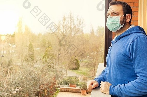 由于冠状病毒covid19大流行，在隔离期间，戴着口罩的年轻高加索人望着家阳台。