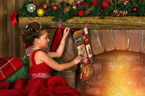 冬天公主-穿着红色连衣裙的可爱小女孩把礼物放进挂在壁炉上的袜子里。欢<strong>迎新</strong>年、圣诞<strong>节</strong>