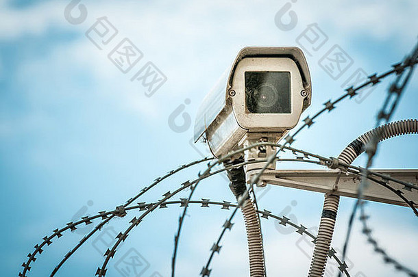 悬挂在监狱铁丝网或其他有蓝天背景的受保护物体之间的安全摄像头的特写镜头。现代方式