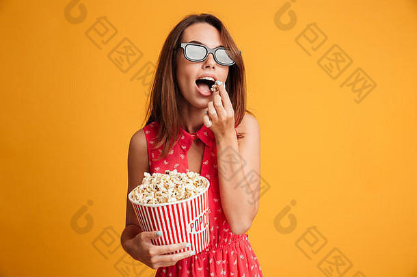 一位戴着3d眼镜、身穿红色连衣裙的年轻黑发美女，一边吃着爆米花，一边看着摄像机，被隔离在黄色背景下