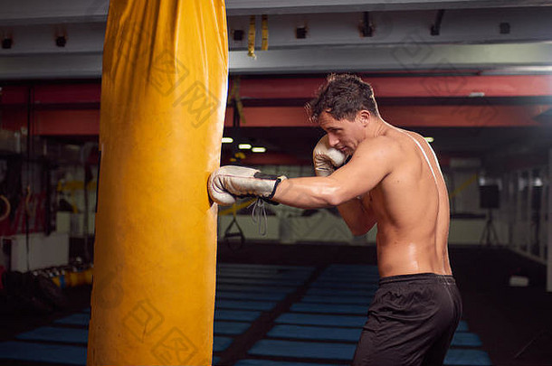 一个年轻的赤膊男子，拳击手用手击打拳击袋，在室内健身房练习，戴拳击手套，上身射击。