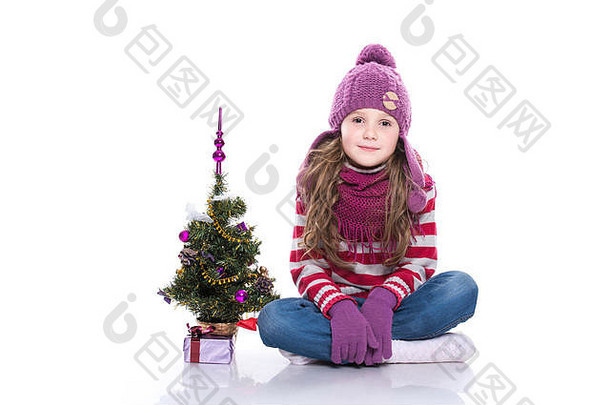 可爱的微笑小女孩，戴着紫色针织围巾和帽子，坐在圣诞树旁，白色背景上的礼物被隔离开来。冬衣和c