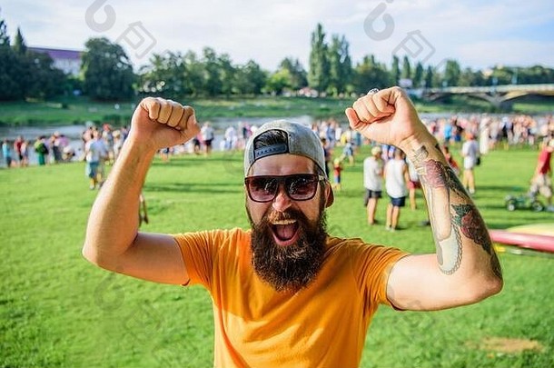<strong>夏日狂欢</strong>节上欢快的歌迷。满脸胡须的潮人在人群前举起拳头绿色的河畔背景。城市活动庆典。嬉皮士戴帽欢庆活动野餐节或节日。